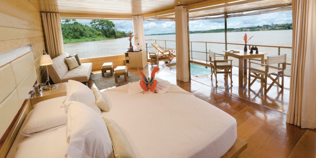 A luxurious suite on the Delfin I cruise ship, featuring a king-size bed, a private plunge pool, and floor-to-ceiling windows with stunning views of the Amazon River.